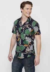 Camisa de botones de manga corta en color marino con patrones de hojas tropicales en verde y beige, fabricada con un tejido ligero. Cuenta con cuello y bolsillo en el pecho.
