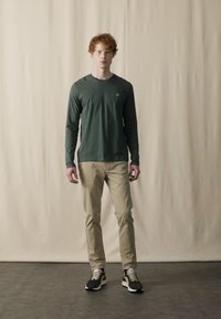 Langærmet grøn skjorte, beige bukser, sorte og hvide sneakers. Model står foran en neutral baggrund, med en slim fit og afslappet stil.