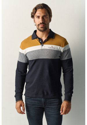 Hombre con pelo corto y castaño y barba, vestido con una camisa polo de manga larga con bloques de color mostaza, blanco, gris y azul marino, de pie frente a una pared lisa.
