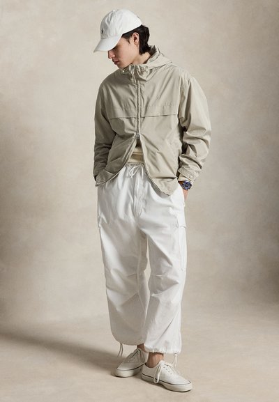 Jeune homme portant une veste à capuche beige, un pantalon cargo blanc, des baskets blanches et une casquette blanche, posant sur un fond neutre.