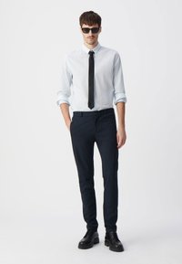 Lichtblauw gestreepte shirt, zwarte stropdas, donkere taps toelopende broek en zwarte schoenen. Model draagt een zonnebril, heeft kort haar en zijn/haar armen langs het lichaam.