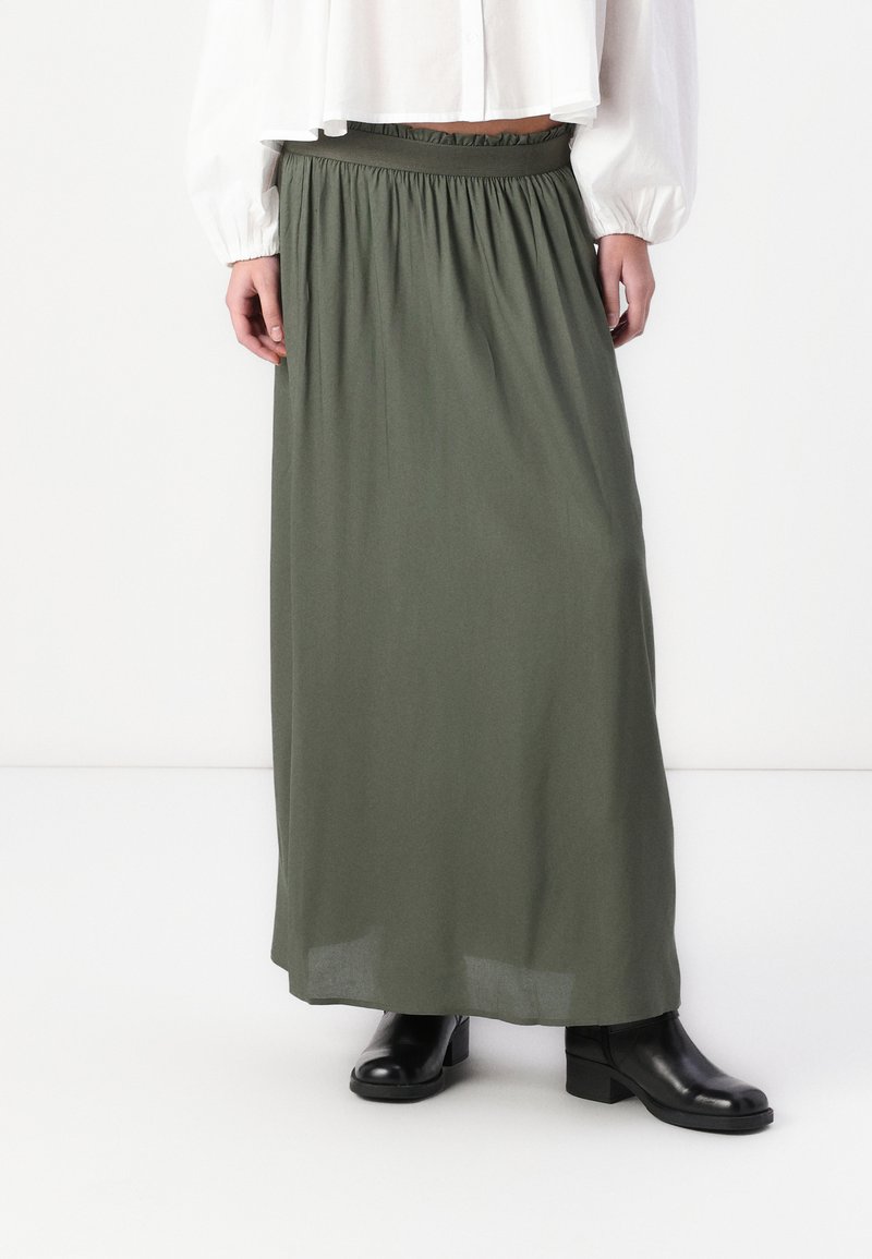 Jupe maxi vert olive avec une taille élastique, fabriquée en tissu fluide. Associée avec des bottines noires et un blouson blanc court.
