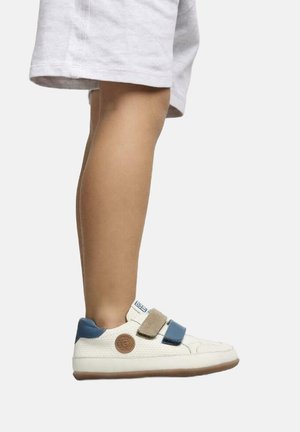 Zapatillas blancas con tiras de velcro azul y beige, parte superior texturizada, parche de logo redondo en el costado, contrastadas por una suela de goma marrón.