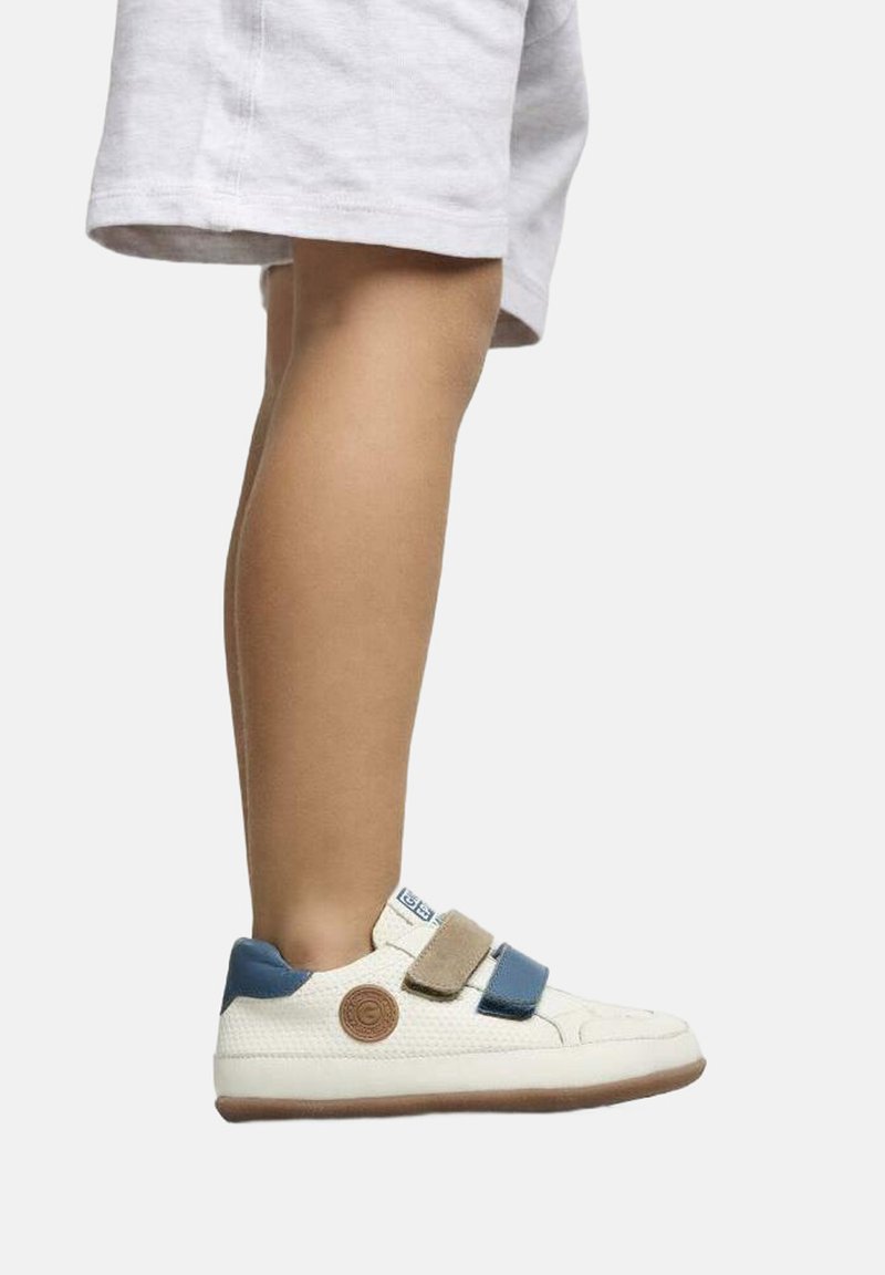Zapatillas blancas con tiras de velcro azul y beige, parte superior texturizada, parche de logo redondo en el costado, contrastadas por una suela de goma marrón.