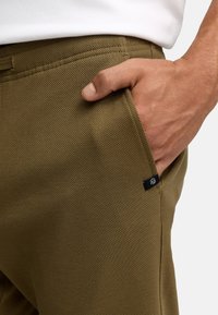 Main d'homme reposant dans la poche avant d'un pantalon en tissu côtelé vert olive avec une petite étiquette noire sur la couture.