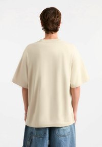 Beżowy t-shirt z bawełny, z krótkimi rękawami, luźnym krojem i zaokrąglonym dekoltem, w zestawie z niebieskimi szortami dżinsowymi z kieszeniami.