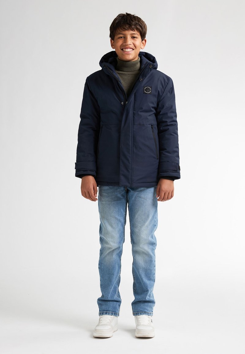 Giacca isolante blu navy con cappuccio, chiusura frontale e due tasche laterali. Indossata sopra un dolcevita verde e abbinata a jeans blu e sneakers bianche.