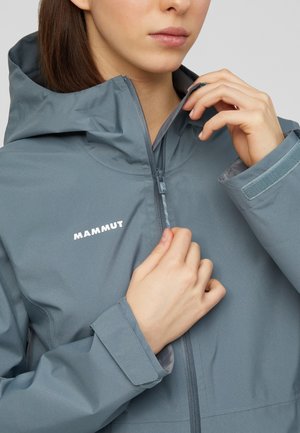 Persoon die een grijze Mammut capuchonjas met klittenbandmanchetten dichtritst, met focus op de bovenste romp en handen.