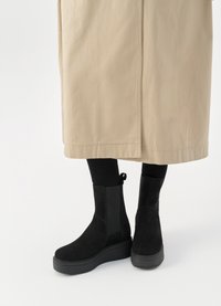 Černé semišové kotníkové boty s tlustou podrážkou, se zasunutými bočními panely a poutky, v kombinaci s dlouhým béžovým svrchním oděvem.