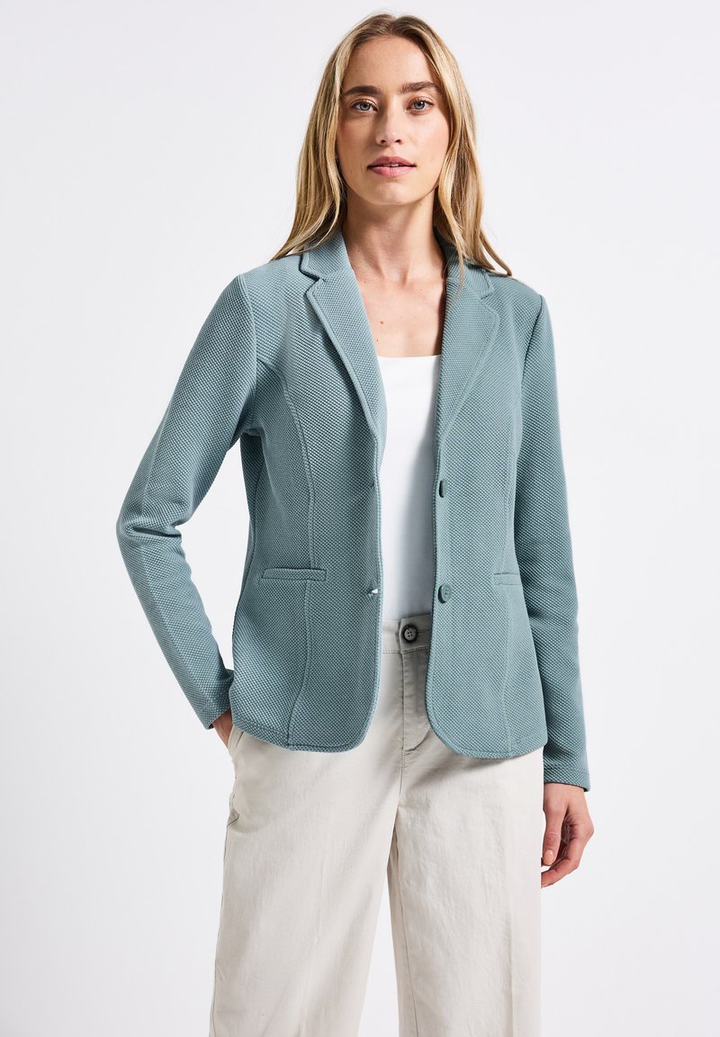 Světle modrý strukturovaný blazer s klopou, dvěma knoflíky a dvěma předními kapsami. Nosený přes bílý top a béžové kalhoty.
