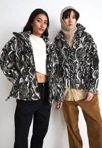Fleece jackets with a bold, geometric black, white, and gray pattern. Zip closures and adjustable hems. One model wears a beige knit hood.