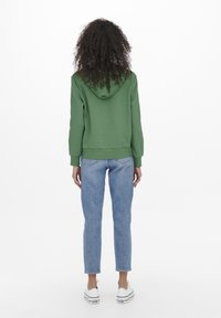 Sudadera verde con capucha, puños y dobladillo acanalados, combinada con vaqueros azules. El atuendo se completa con zapatillas blancas, visto desde atrás.