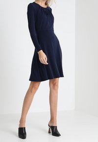 Marinblå långärmad klänning med en åtsittande bodice, flödande kjol och draperad halsringning. Bärs med svarta klackmules.