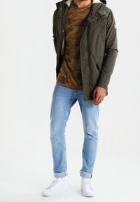 Olijfgroene jas met een witte fleece voering, camouflage t-shirt, lichtblauwe jeans en witte sneakers. De jas heeft een capuchon en zakken.