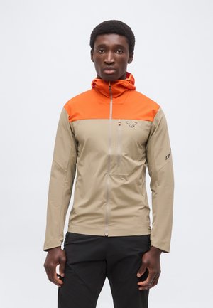 Άνδρας με μπεζ και πορτοκαλί ζακέτα με φερμουάρ και κουκούλα και μαύρο παντελόνι, κοιτάζει μπροστά σε ένα απλό φόντο.