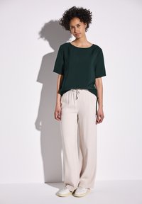 Tmavě zelené tričko s kulatým výstřihem a krátkým rukávem, v kombinaci se světle béžovými širokými kalhotami a bílými teniskami. Hladké textury, ležérní design.