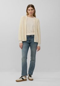 Romige textuur cardigan over een gestreept beige top, gecombineerd met vervaagde blauwe jeans en sneakers met zwarte accenten en tanzolen.