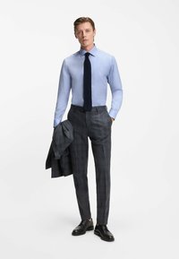 Lichtblauw overhemd, marineblauwe stropdas, grijs geruite pantalon en zwarte leren schoenen. Model houdt een bijpassende grijze blazer over zijn arm.