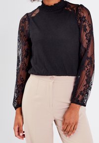 Haut noir avec corsage texturé et manches longues en dentelle noire transparente, doté d'un col froncé. Associé à un pantalon beige taille haute.