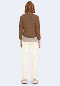 Brauner gestrickter Pullover mit runden Ausschnitt, der einen helleren gerippten Saum hat. Kombiniert mit weißen lässigen Hosen und cremefarbener Schuhbekleidung.