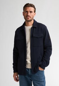 Marineblauwe geweven overhemdjack met twee voorzakken, drukknoopsluiting en een relaxte pasvorm. Draag het over een lichtbeige trui en blauwe jeans.