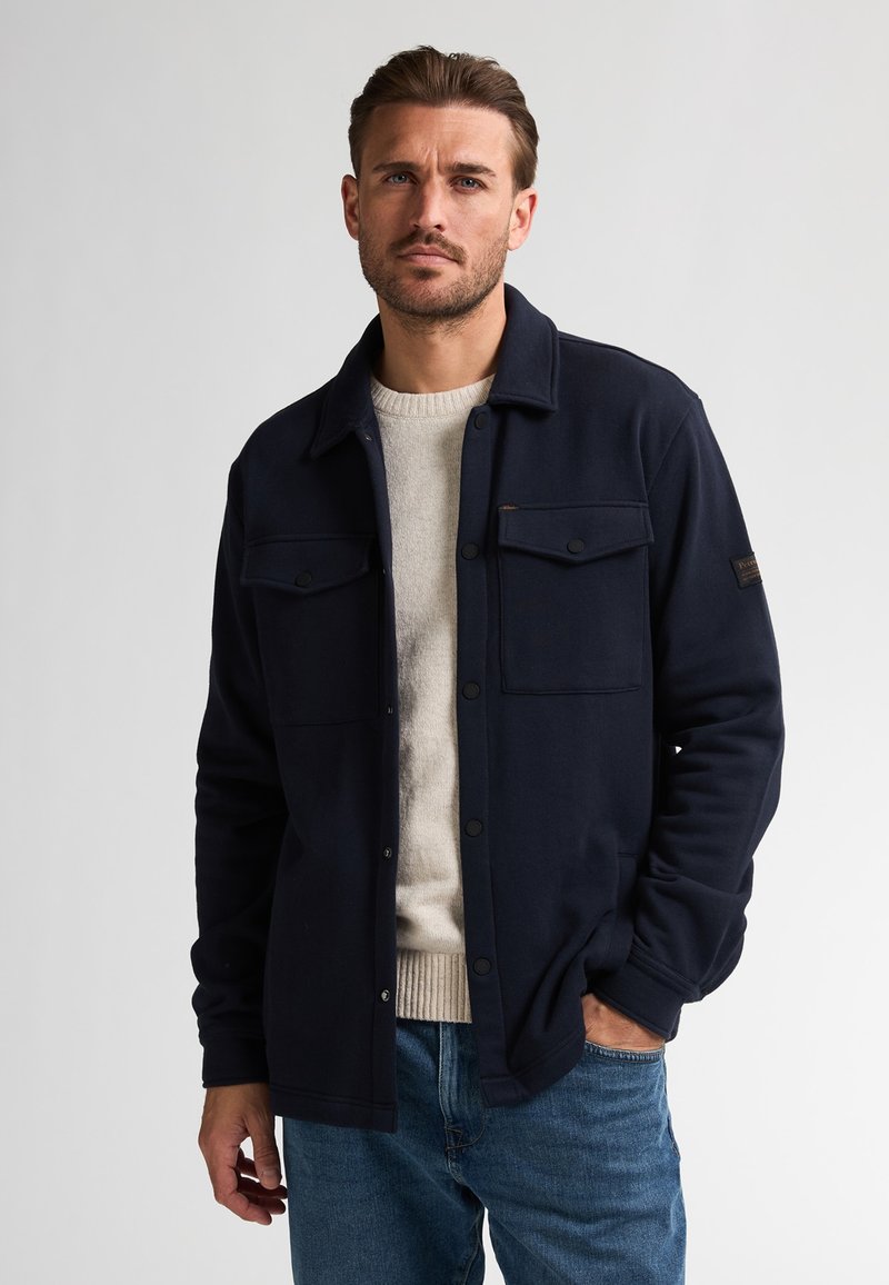 Marineblauwe geweven overhemdjack met twee voorzakken, drukknoopsluiting en een relaxte pasvorm. Draag het over een lichtbeige trui en blauwe jeans.