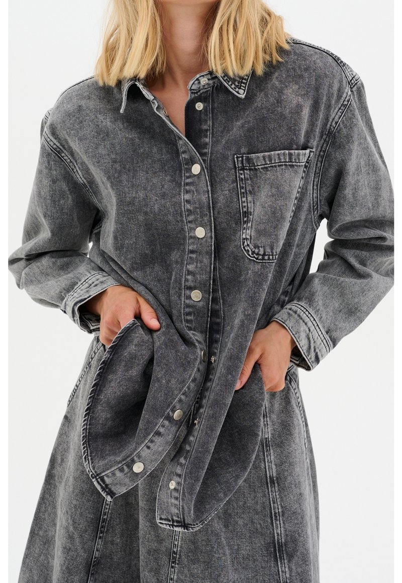 Personne blonde portant une veste en jean noire délavée oversize avec des boutons-pression, les mains tirant un côté de la veste près des poches.