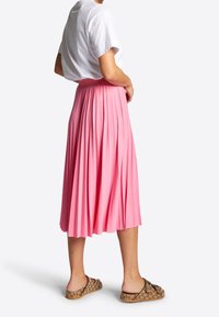 Femme portant une chemise blanche à manches courtes, une jupe midi plissée rose et des sandales à enfiler beiges de créateur avec des doubles boucles, se tenant sur fond blanc.