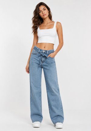 Kvinde iført en hvid crop top og højtaljede, bredbenede blå jeans med bindebælte, står med den ene hånd i lommen.