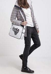 Borsa a spalla Gola bianca con logo e dettagli neri, abbinata a un giubbotto imbottito grigio, top bianco e jeans neri. Le scarpe nere completano il look.