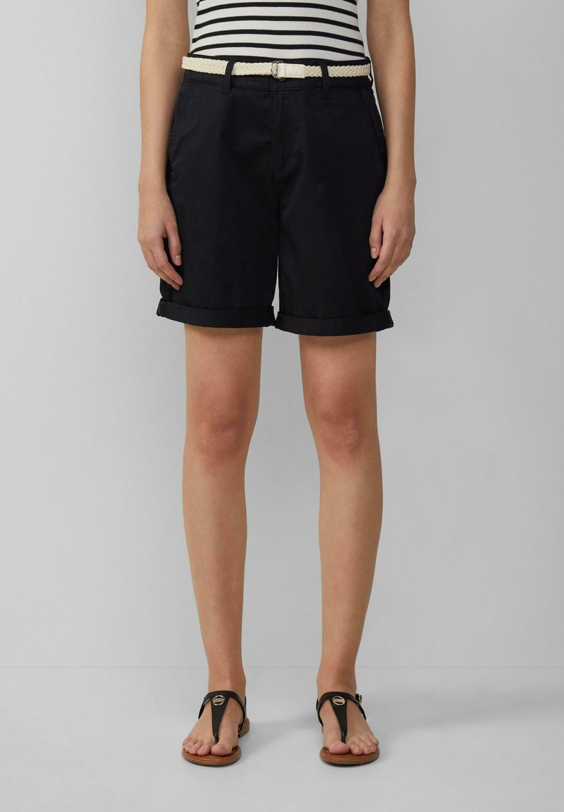 Shorts noirs à ourlet roulé avec poches latérales, associés à une ceinture tressée blanche. Portés avec des tongs noires mettant en valeur un accent métallique rond.