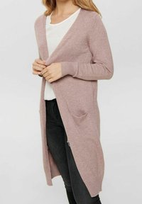 Cardigan lungo aperto color rosa chiaro, realizzato in materiale morbido. Presenta polsini a costine e due tasche frontali, abbinato a una maglietta bianca e jeans neri.