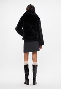 Casaco de pelúcia preto com uma gola larga, mangas compridas e uma bainha ligeiramente cropped, acompanhado por uma saia preta e botas brilhantes até ao joelho.