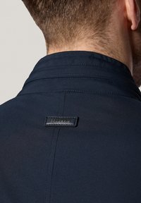 Granatowa kurtka z wyraźnym kołnierzem, gładkim materiałem oraz naszywką z logo na plecach z czarnym skórzanym akcentem.