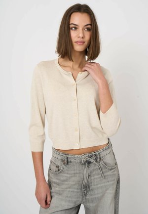 Kobieta o brązowych włosach do ramion, ubrana w beżowy, zapinany na guziki kardigan i wysokie szare dżinsy, spoglądająca w bok z ręką przy szyi.