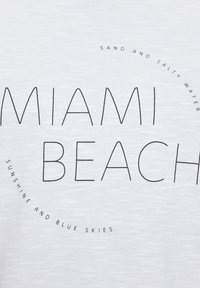 White cotton t-shirt featuring black text with "MIAMI BEACH" prominently displayed, surrounded by circular phrases in a modern font.