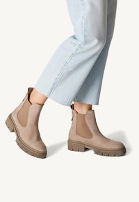 Bottes chelsea en suède beige avec des panneaux latéraux élastiques, semelles en caoutchouc texturé et tirettes, présentées avec un jean cropped bleu clair.