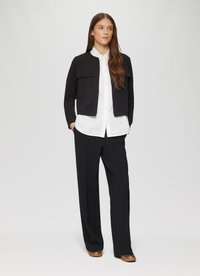 Veste cropped noire avec deux poches avant portée sur une chemise blanche à boutons, associée à un pantalon large noir et des chaussures marron.