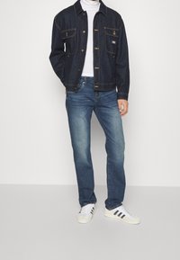 Person som bär en mörk jeansjacka, vit polotröja, blå jeans och vita sneakers med svarta ränder, står mot en enfärgad vägg.