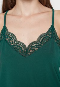 Camisola teal con delicate rifiniture in pizzo lungo un scollo a V e spalline regolabili. La texture liscia si abbina ai dettagli intricati in pizzo.