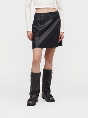 Femme portant une mini-jupe en cuir noire, des bottes en cuir noires montant jusqu’au genou et un top blanc à manches longues coupé, debout devant un fond uni.