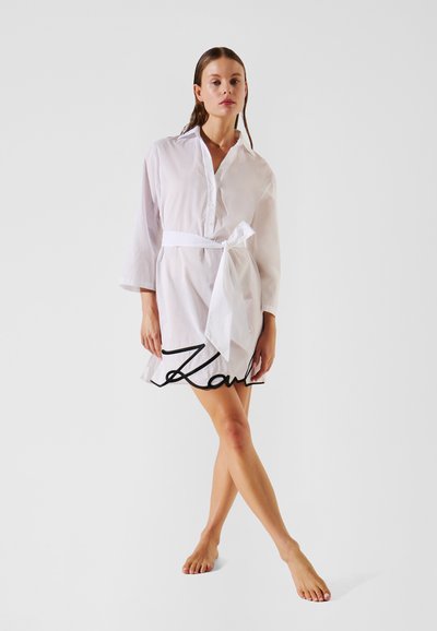 Robe blanche à boutons avec des manches longues et une ceinture à nouer à la taille, en tissu léger et transparent, présentant une silhouette droite et fluide.