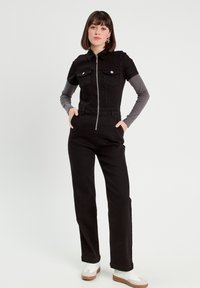 Salopette in denim nero con maniche corte, zip sul davanti, due tasche sul petto, abbinata a una camicia grigia a maniche lunghe e sneakers bianche. Texture liscia.