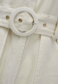 Beige stofbælte med rund stofbetrukket spænde og metalnål trukket gennem metaløje på matchende beige beklædning.