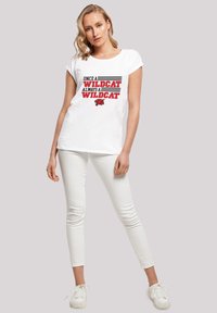 Hvit kortermet t-skjorte med "ONCE A WILDCAT ALWAYS A WILDCAT" trykket i rødt og svart, med grafikk av en villkatt. Kombinert med hvite bukser og sko.