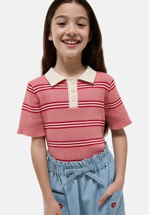 Fille souriante portant un polo rose côtelé à manches courtes avec des rayures rouges et blanches et un pantalon taille paperbag bleu clair avec broderies florales.