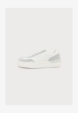 Zapatilla deportiva blanca de caña baja con detalles en ante gris claro y suela blanca gruesa, mostrada desde un lateral sobre un fondo claro y liso.