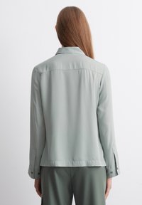 Lys grøn bluse med en glat tekstur, der har en skræddersyet ryg, krave og knapmanchetter. Designet er minimalistisk uden synlige mønstre.