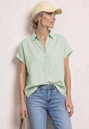 Mujer con una gorra amarillo pálido, camisa de manga corta verde claro con botones y vaqueros azules claros de talle alto, de pie contra una pared clara.