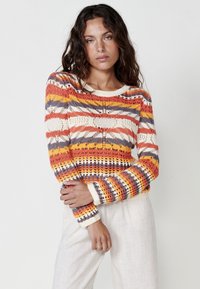 Suéter de punto multicolor con un patrón en forma de flecha en naranja, amarillo, púrpura y blanco. Mangas largas con puños y cuello acanalados.